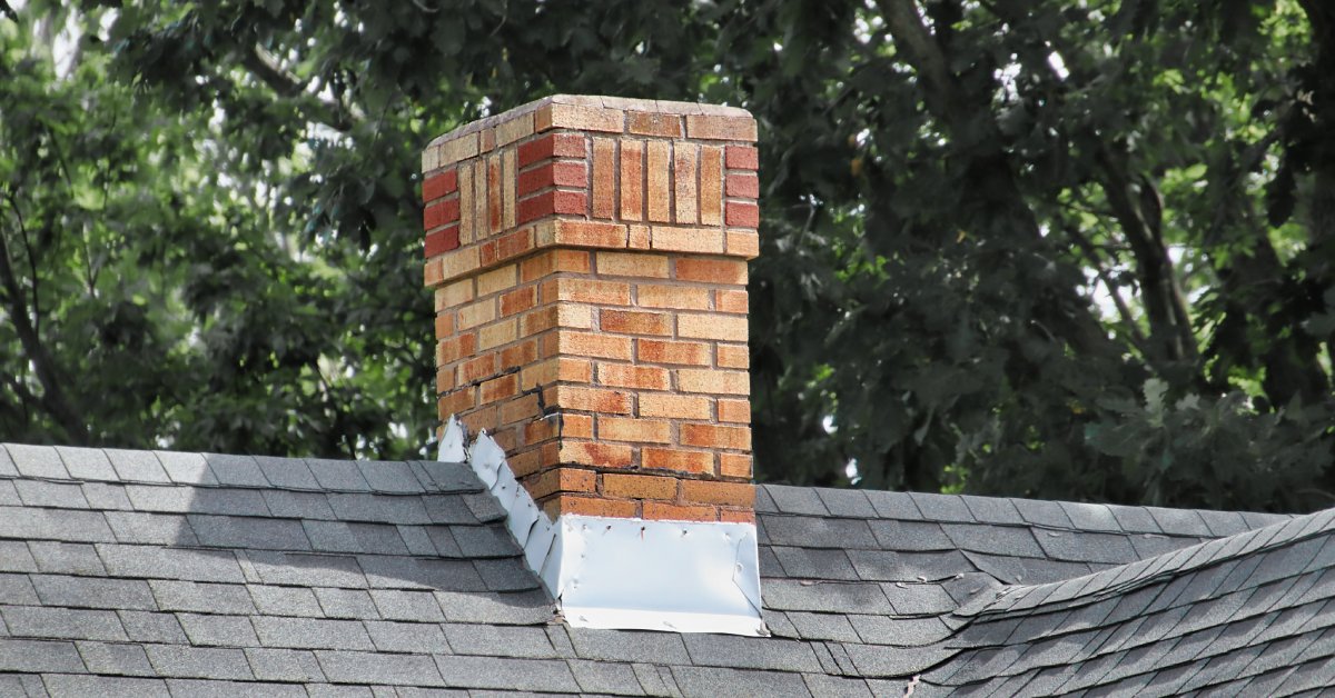 A brick chimney with a metal base stands on top of a home with a gray shingled roof near tall trees.