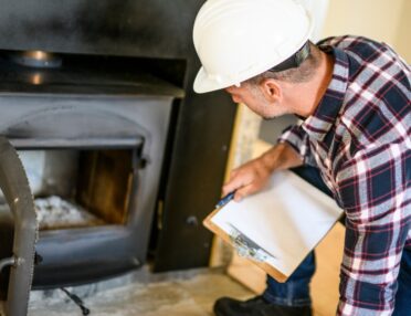 An inspector wears a hard hat and holds a clipboard in one hand while kneeling to look at a fireplace.