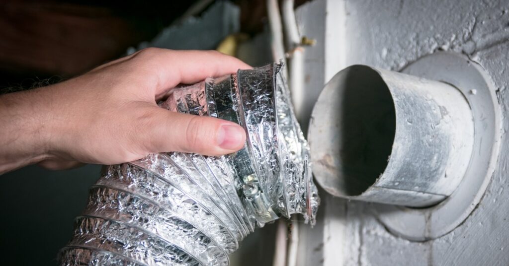 A person holding an aluminum dryer vent hose in their hand, disconnecting it from the exit point in their wall.
