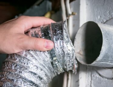 A person holding an aluminum dryer vent hose in their hand, disconnecting it from the exit point in their wall.