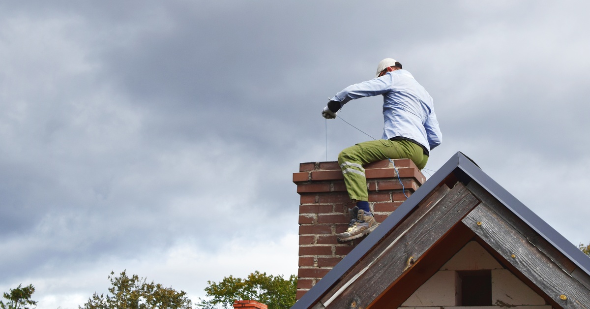 A worker sitting on a chimney and using a tool to clean the inside. There are some dark and stormy clouds in the background.