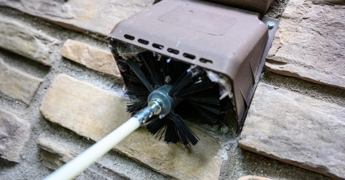 A person uses a large spinning brush to clean the exterior dryer vent on their home. The vent comes out of a stone wall.