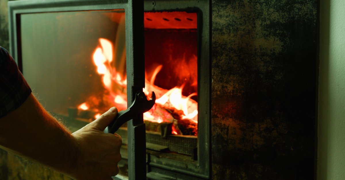 A person opens the glass door on a fireplace that has some large red flames burning a pile of wood.