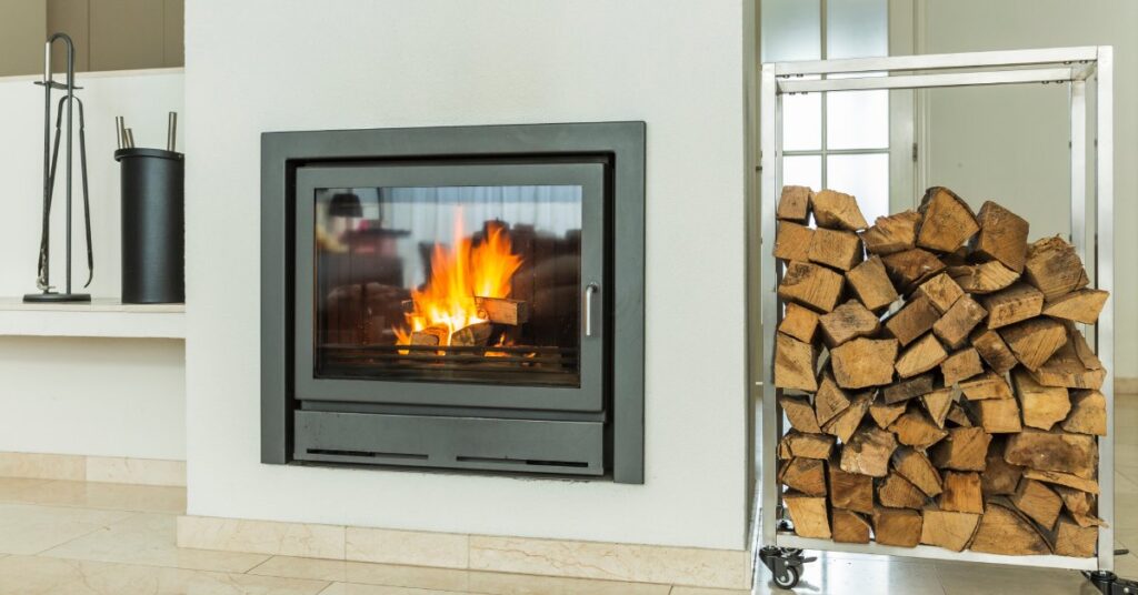 A close-up of a wood-burning fireplace with a glass door in front of it and a stack of firewood to the right side.