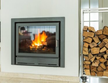A close-up of a wood-burning fireplace with a glass door in front of it and a stack of firewood to the right side.