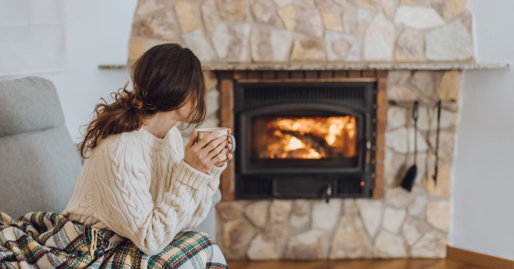 A woman sitting on the edge of her chair, holding a mug, while looking at her fireplace. The fire is roaring.