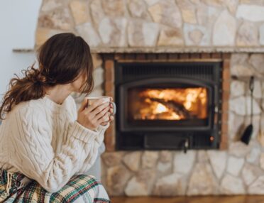 A woman sitting on the edge of her chair, holding a mug, while looking at her fireplace. The fire is roaring.